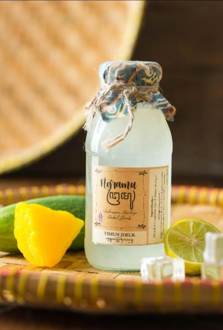 Botol kaca Timun Jeruk berlabel Ngramu bertutup kain batik biru di nampan anyaman, dengan timun dan jeruk nipis