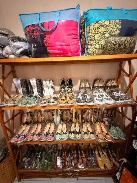 Rak kayu berisi deretan sepatu dan slip on bermotif batik dalam plastik, dengan dua tas besar di atasnya