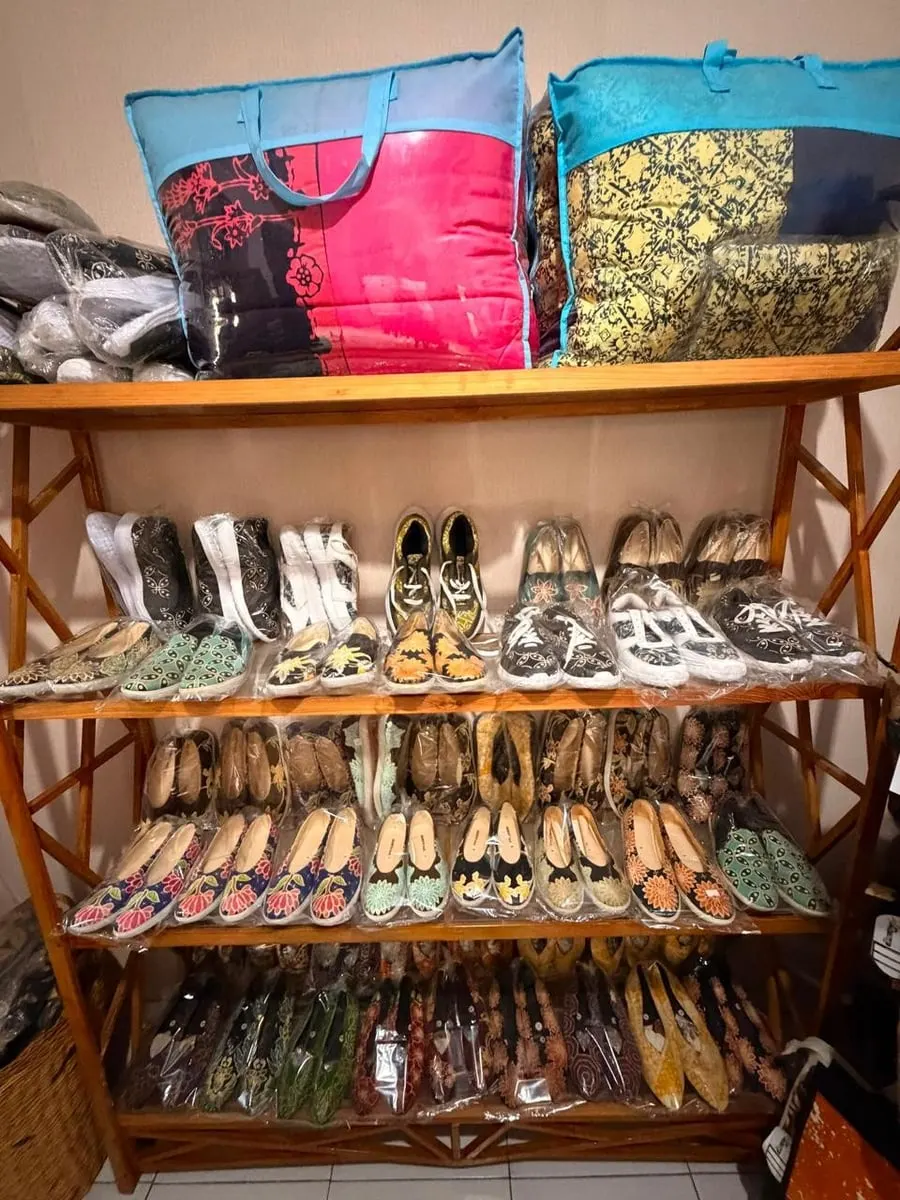 Rak kayu berisi deretan sepatu dan slip on bermotif batik dalam plastik, dengan dua tas besar di atasnya