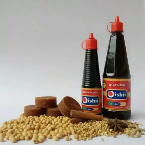 Dua botol Kecap Manis Oishii berlabel merah di samping gula merah, kayu manis, kedelai, dan ketumbar