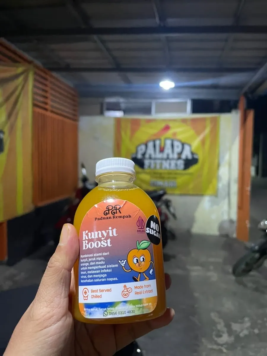 Tangan memegang botol Kunyit Boost berisi minuman oranye di depan spanduk Palapa Fitness