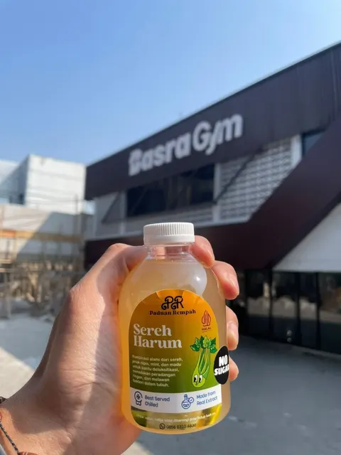 Tangan memegang botol Sereh Harum berisi minuman kuning di depan sebuah gedung gym di bawah langit biru