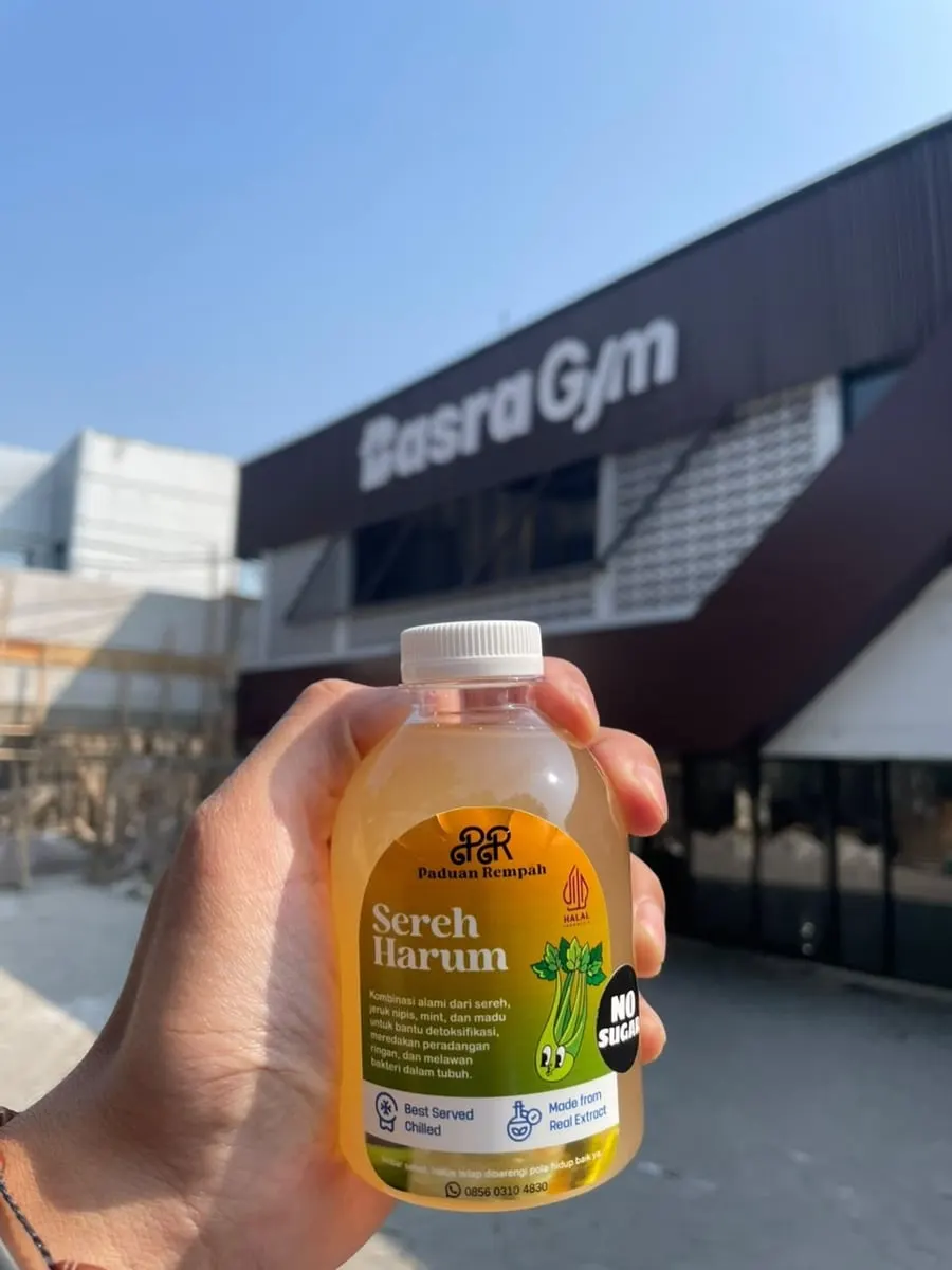 Tangan memegang botol Sereh Harum berisi minuman kuning di depan sebuah gedung gym di bawah langit biru