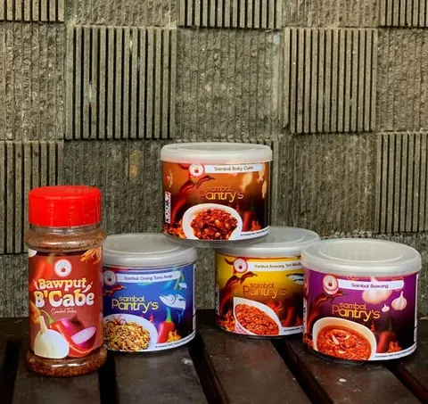 Empat tabung sambal Pantry's warna-warni dan satu botol Bawput B'Cabe bertutup merah di atas bangku kayu