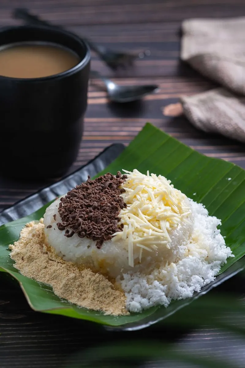 Ketan putih di atas daun pisang dengan taburan meses cokelat, keju parut dan bubuk kedelai, di samping cangkir kopi