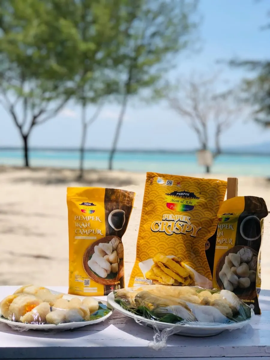 Kemasan kuning Pempek Ikan Campur dan Pempek Crispy Mom & Khi dengan dua piring pempek di meja tepi pantai