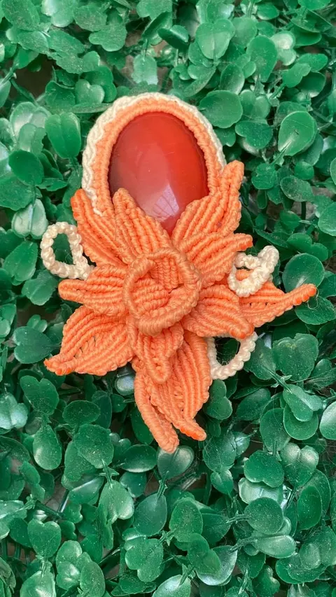 Aksesori macrame oranye berbentuk bunga dengan batu merah oval di atas latar daun hijau