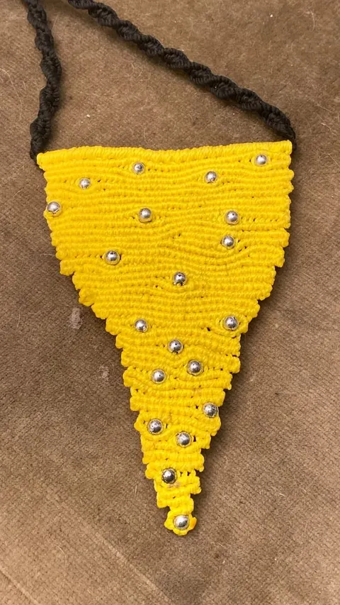 Kalung macrame kuning berbentuk segitiga bertabur manik perak dengan tali hitam di atas latar cokelat