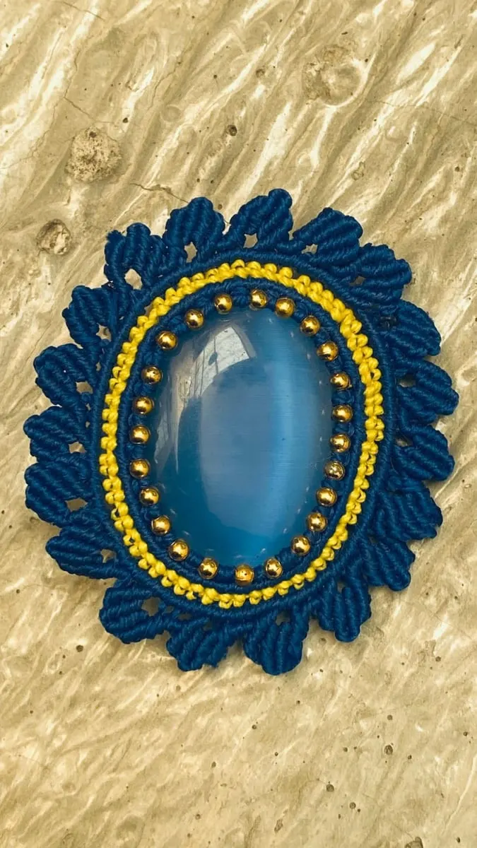 Aksesori macrame biru tua berbentuk oval dengan batu biru di tengah dan manik emas di atas latar krem