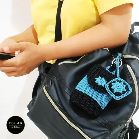 Pouch rajut hitam biru dan gantungan granny square biru tergantung di tas ransel hitam yang dipakai seseorang