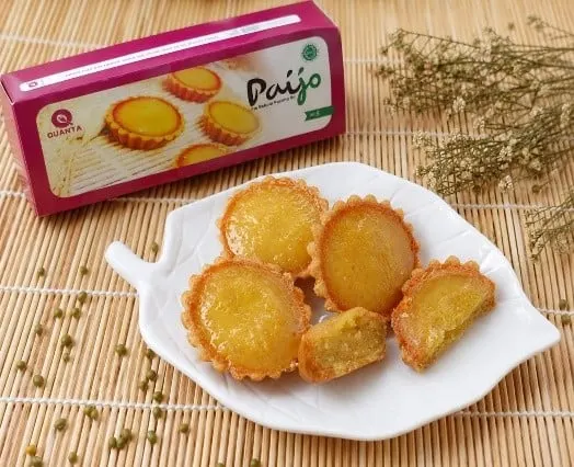 Kotak merah muda Paijo dan piring daun putih berisi pie bakpia kacang hijau di atas tikar bambu