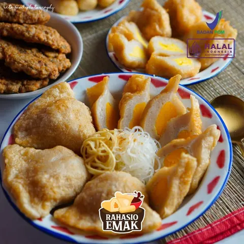 Piring enamel berisi pempek goreng, potongan pempek isi telur, mi kuning, dan bihun, dengan pempek lain di belakang