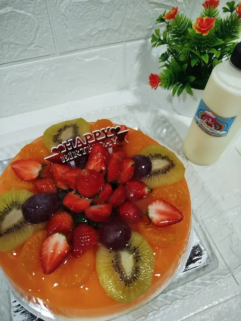 Puding buah oranye berhias stroberi, kiwi, jeruk dan anggur dengan hiasan Happy Birthday dan botol berlabel