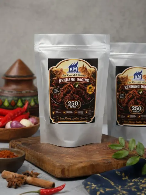 Dua standing pouch perak berlabel Rendang Daging 250 gram di atas talenan kayu dengan cabai dan rempah