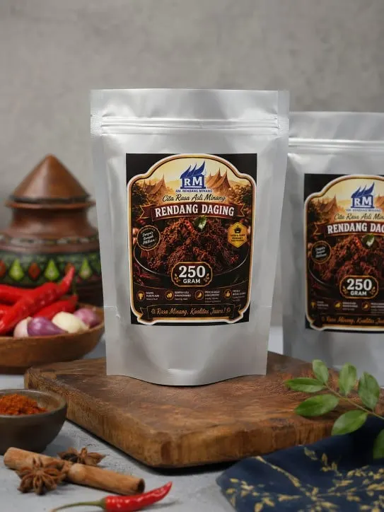 Dua standing pouch perak berlabel Rendang Daging 250 gram di atas talenan kayu dengan cabai dan rempah