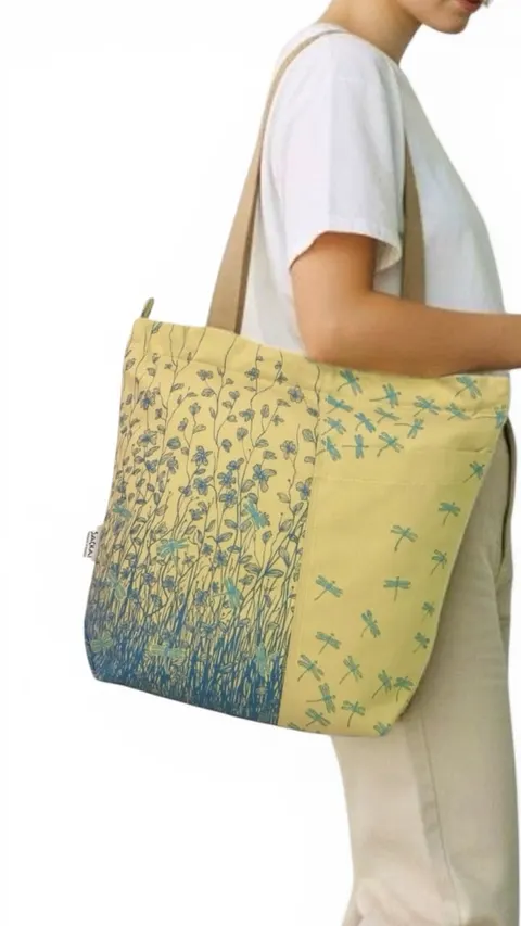 Model membawa tote bag kain kuning bercetak ilustrasi bunga dan capung biru di bahu, berlatar putih
