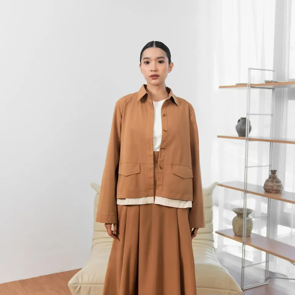 Model memakai jaket kemeja cokelat berkantong, kaus putih dan rok lebar cokelat di ruang dengan rak