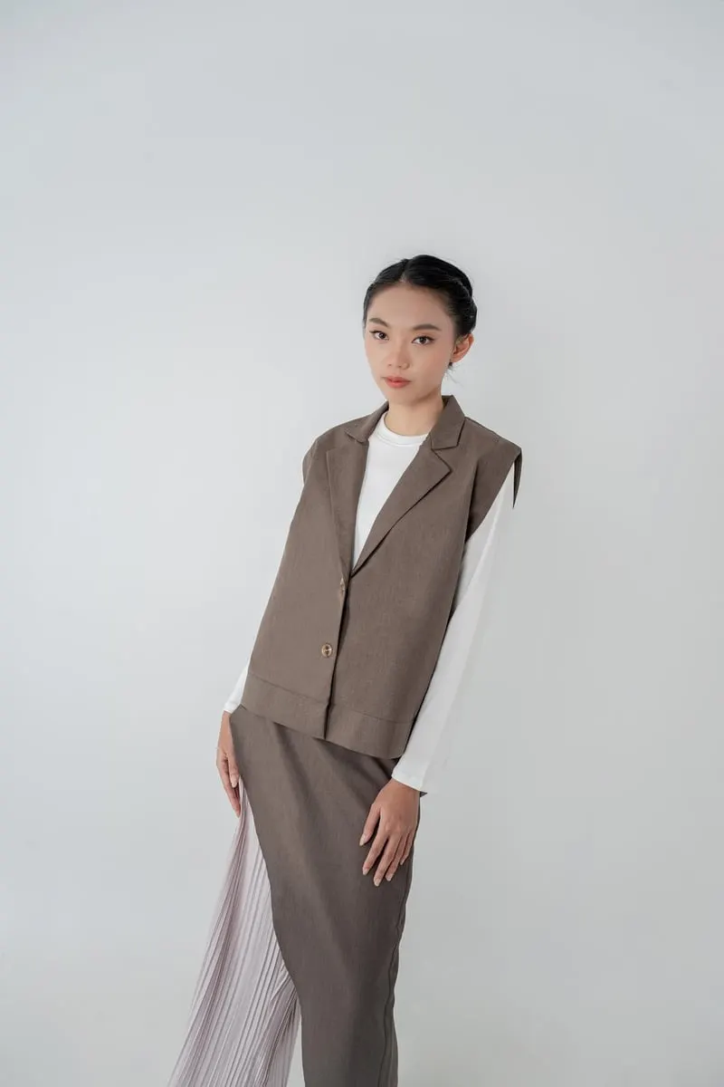 Model memakai vest cokelat tanpa lengan di atas atasan putih dan rok plisket, berlatar abu-abu
