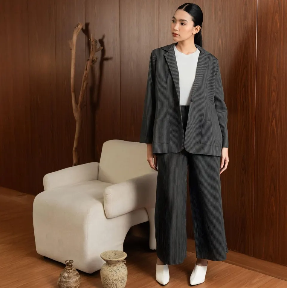 Model memakai setelan blazer dan celana panjang abu tua bergaris dengan atasan putih, di ruang berpanel kayu