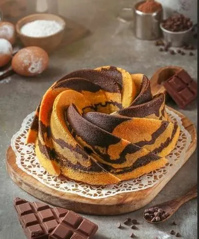 Kue marmer bundar bermotif oranye dan cokelat di atas papan kayu dengan potongan cokelat batangan
