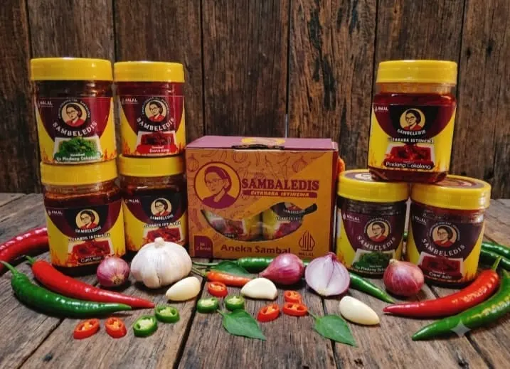 Toples sambal Sambeledis bertutup kuning dan kotak Aneka Sambal di meja kayu, dikelilingi cabai, bawang merah dan putih