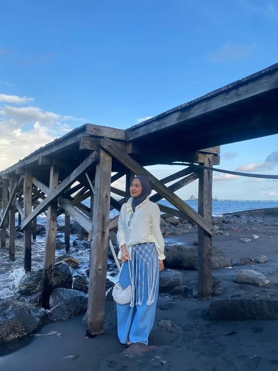 Perempuan berhijab di bawah dermaga kayu di pantai, membawa tas macrame putih dan memakai rumbai macrame