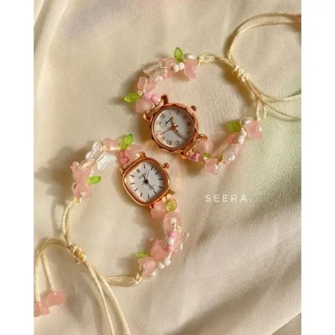 Dua jam tangan berbingkai rose gold dengan tali manik bunga merah muda dan daun hijau di atas kain krem