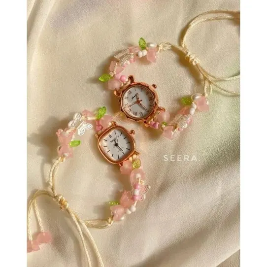 Dua jam tangan berbingkai rose gold dengan tali manik bunga merah muda dan daun hijau di atas kain krem