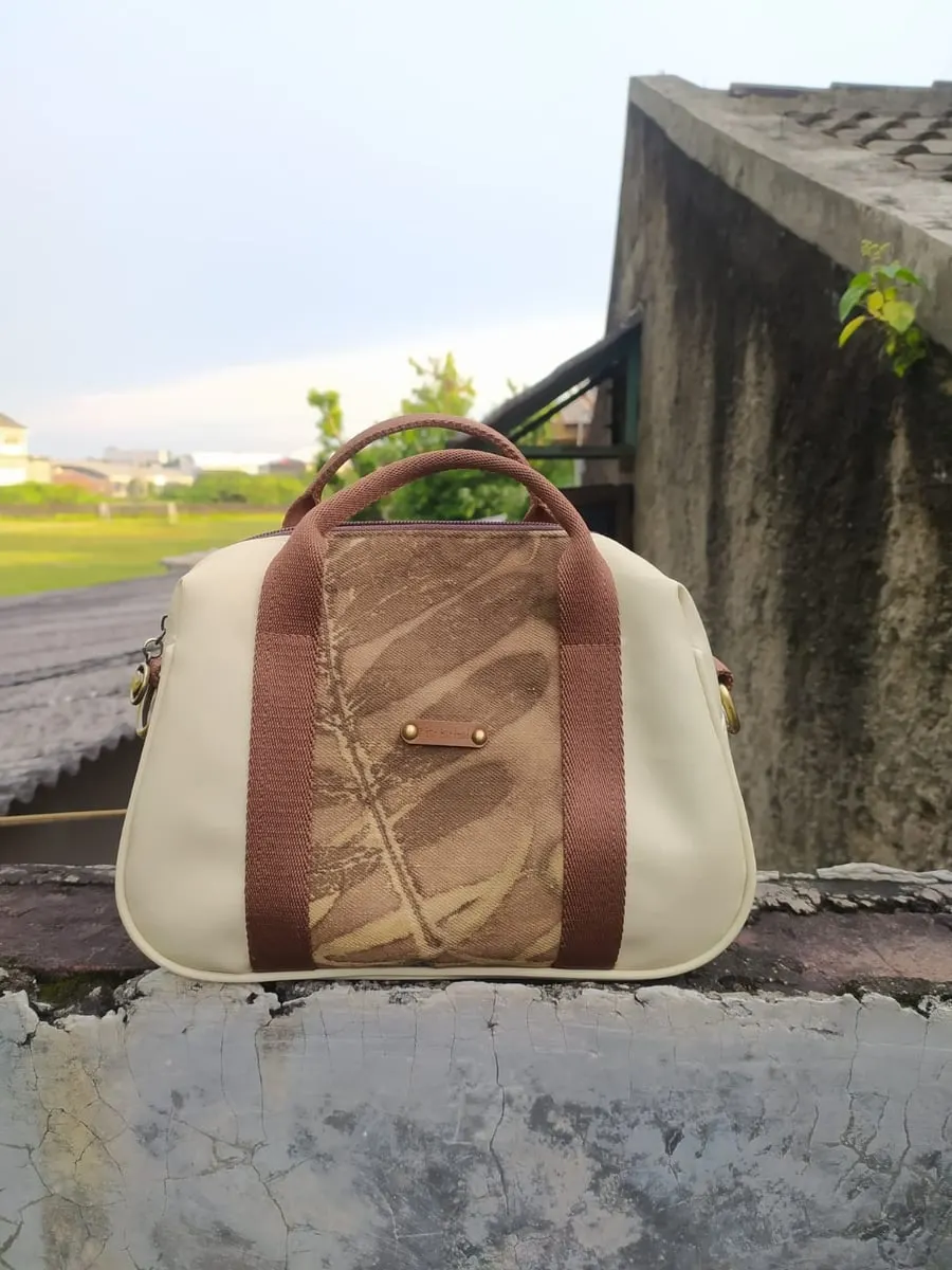 Tas tangan krem dengan panel ecoprint daun cokelat dan tali pegangan cokelat di atas tembok beton