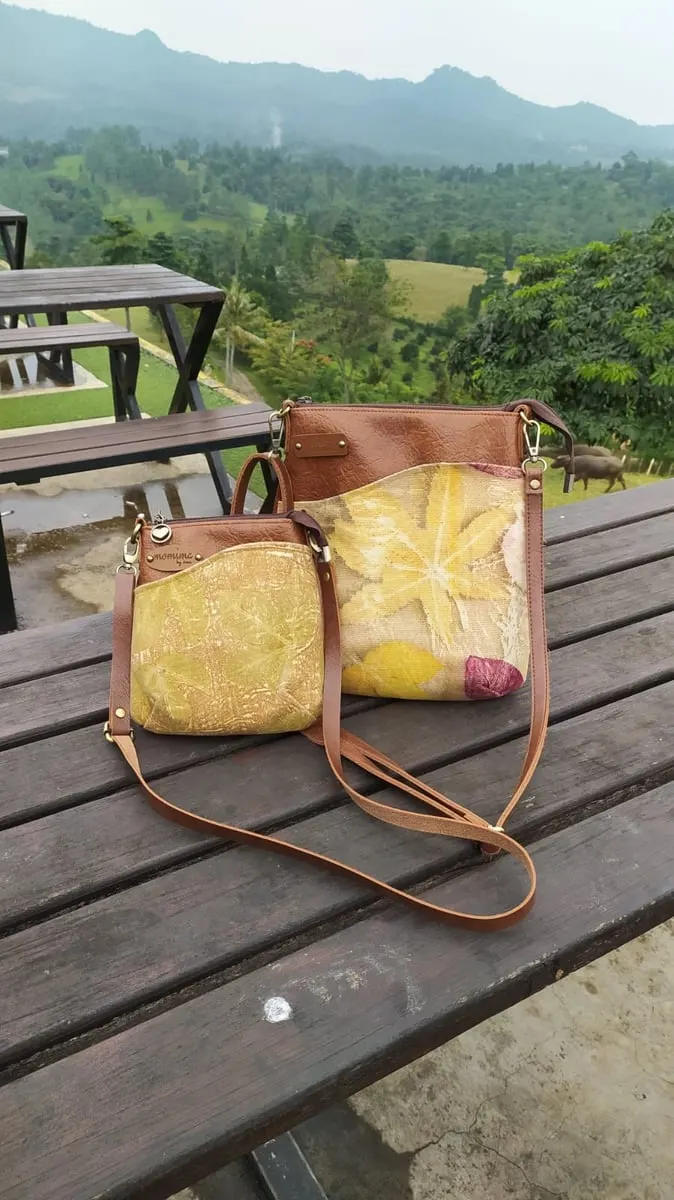 Dua tas selempang bermotif ecoprint daun kuning dengan aksen kulit cokelat di atas meja kayu berlatar perbukitan