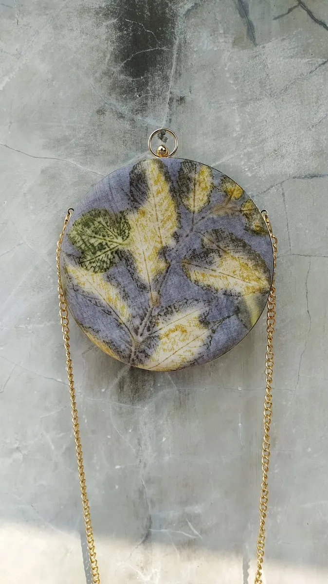 Tas bulat bermotif ecoprint daun kuning di atas warna ungu keabuan dengan tali rantai emas di dinding beton
