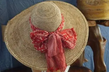 Topi lebar rajutan serat alami berwarna krem dengan pita kain merah bermotif di atas penyangga kayu