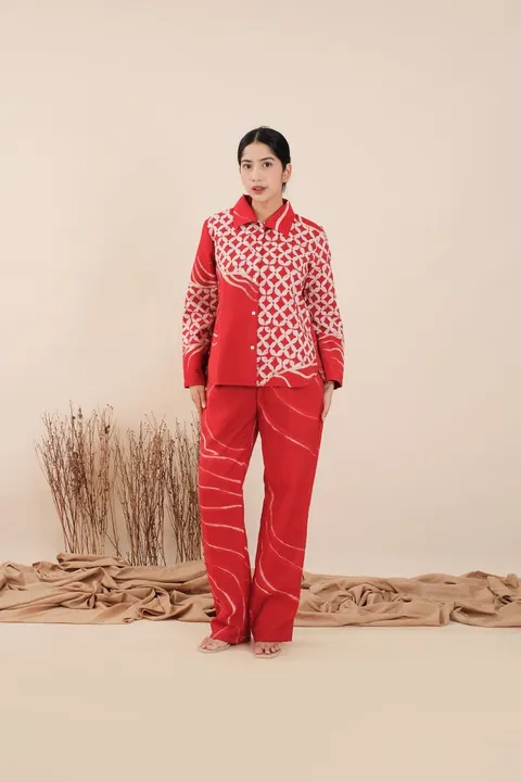 Model memakai kemeja batik merah bermotif putih dan celana merah panjang di studio berlatar krem