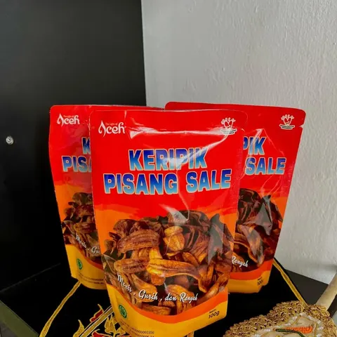 Tiga kemasan pouch merah Keripik Pisang Sale berisi keripik pisang cokelat, di atas kain hitam bersulam emas