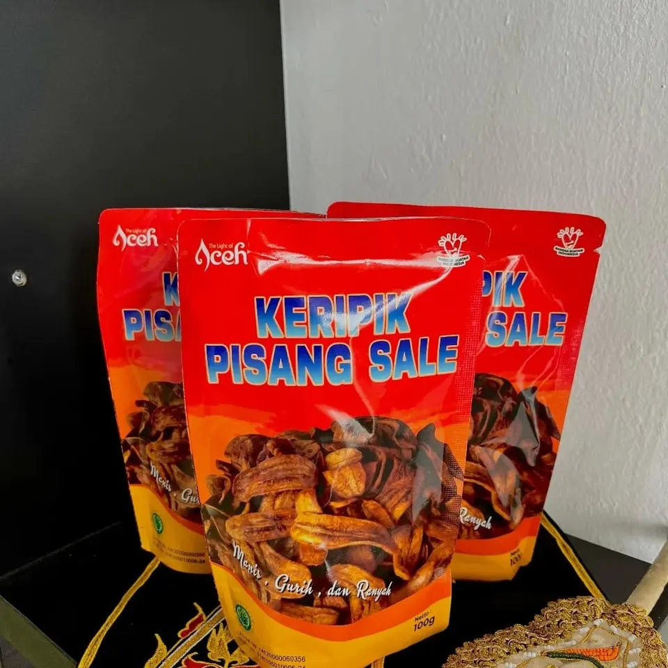 Tiga kemasan pouch merah Keripik Pisang Sale berisi keripik pisang cokelat, di atas kain hitam bersulam emas