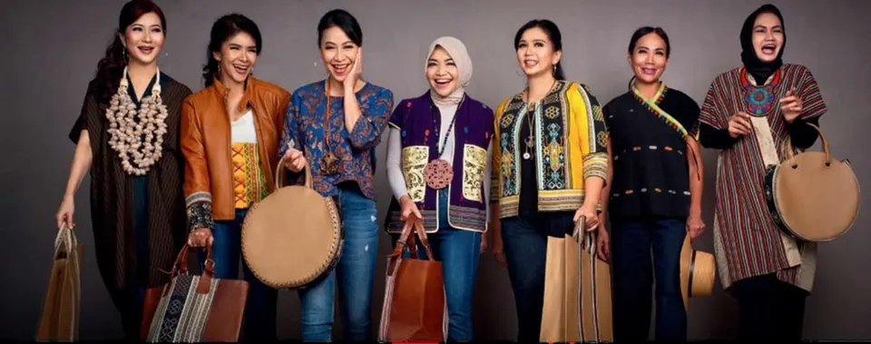 Tujuh perempuan memakai atasan dan rompi bermotif tenun sambil membawa tas kulit dan tas bulat