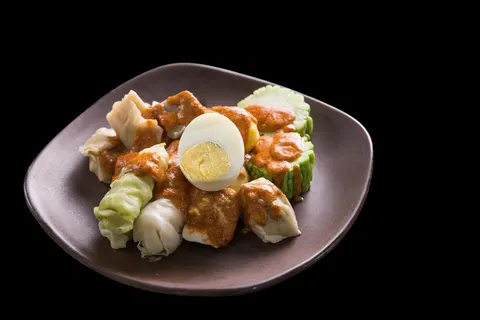 Siomay, kol dan pare bersaus kacang dengan setengah telur rebus di piring keabuan berlatar hitam