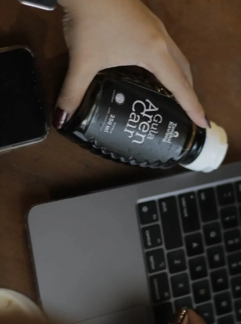 Tangan memegang botol hitam Gula Aren Cair di atas meja dekat laptop dan ponsel