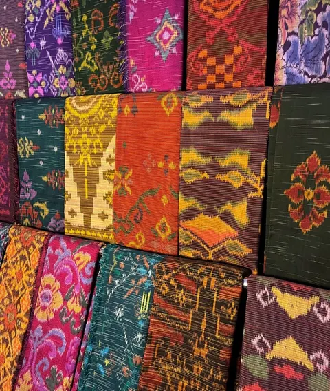 Tumpukan kain tenun endek berwarna merah muda, jingga, hijau dan ungu dengan motif geometris dan bunga