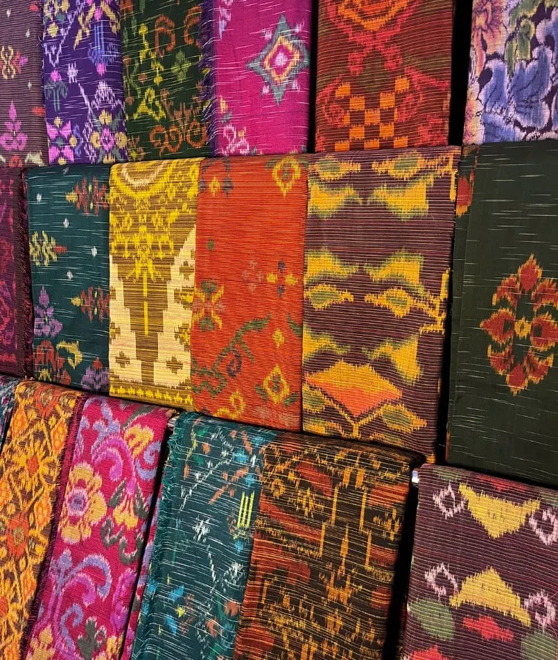 Tumpukan kain tenun endek berwarna merah muda, jingga, hijau dan ungu dengan motif geometris dan bunga