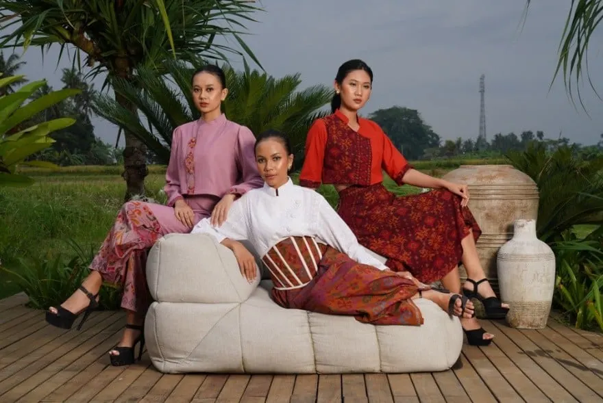 Tiga model mengenakan atasan dan rok berbahan tenun, duduk di sofa putih di dek kayu dekat sawah