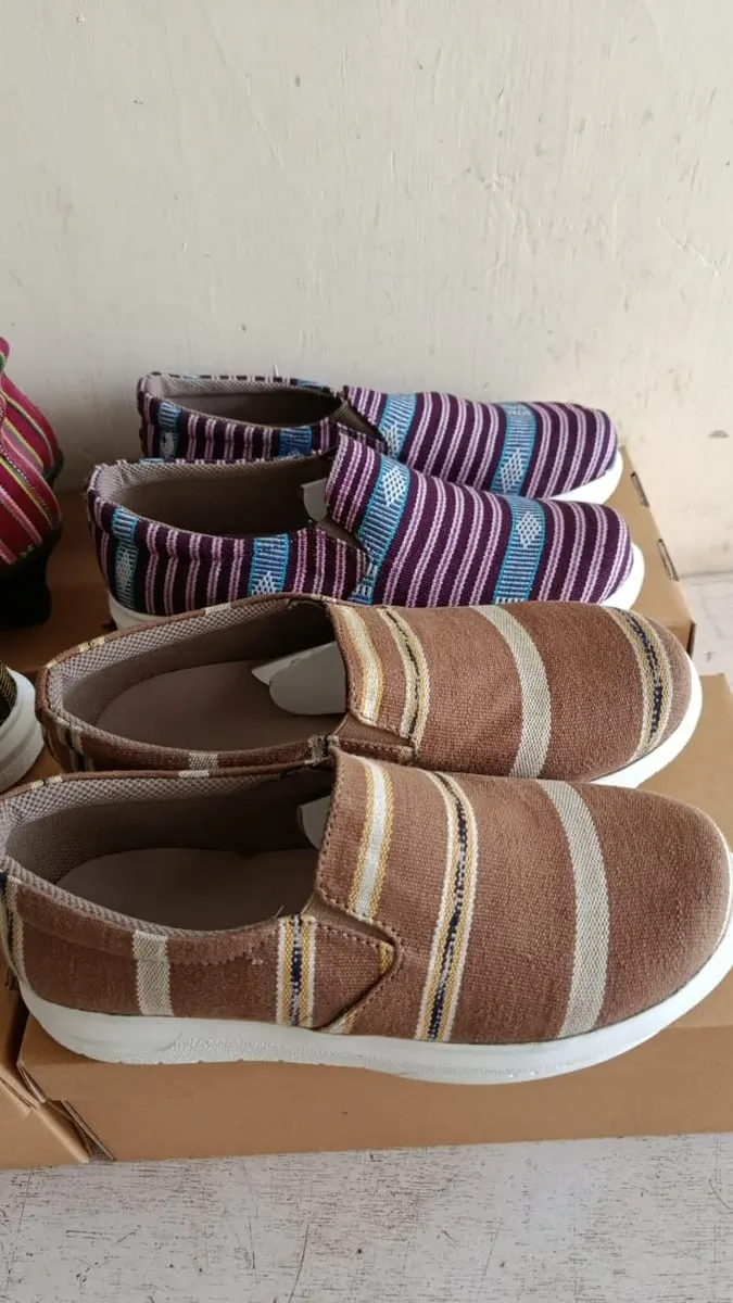 Dua pasang sepatu slip-on berbahan tenun, ungu bergaris dan cokelat bergaris, di atas kotak kardus