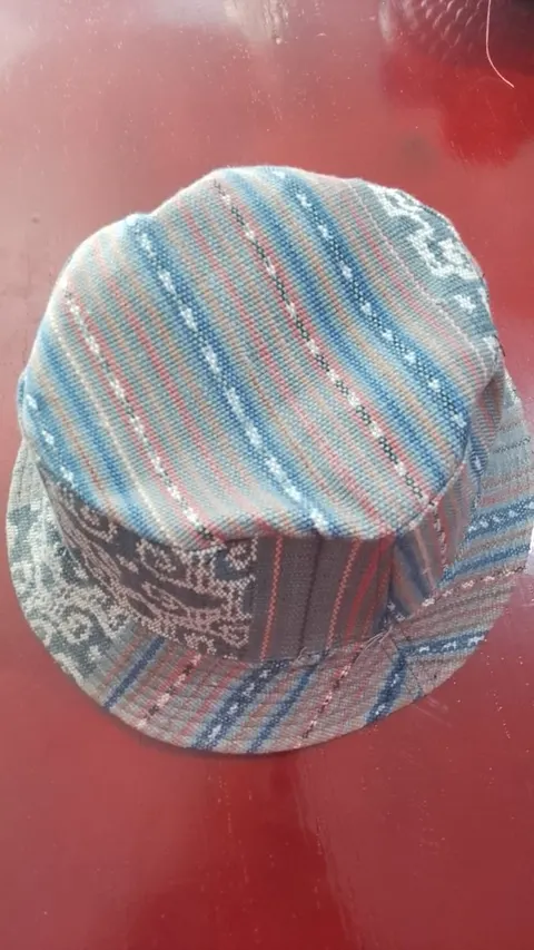 Topi bucket dari kain tenun bergaris biru, merah muda dan abu-abu di atas meja merah