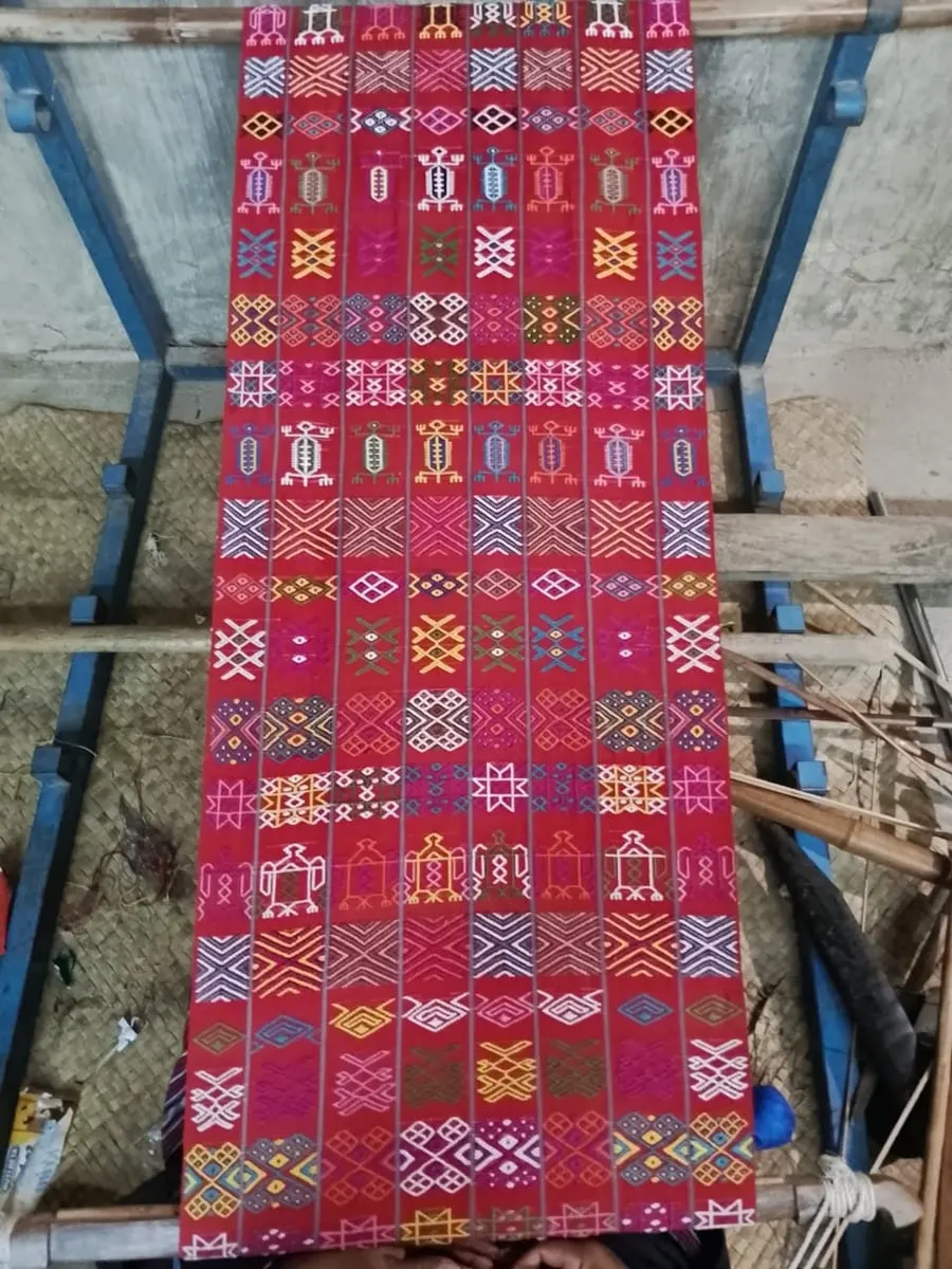 Kain tenun merah bermotif geometris warna-warni terbentang di atas alat tenun kayu