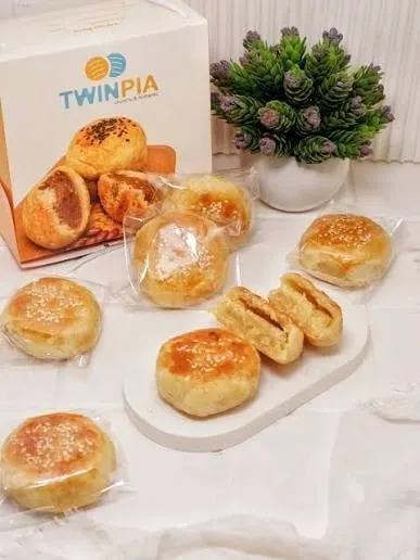 Bakpia dalam plastik satuan di depan kotak putih Twinpia, dua bakpia dibelah memperlihatkan isian