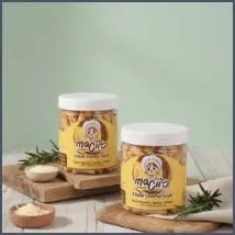 Dua toples plastik bertutup putih berisi stik keju Maciis di atas talenan kayu dengan ranting daun hijau