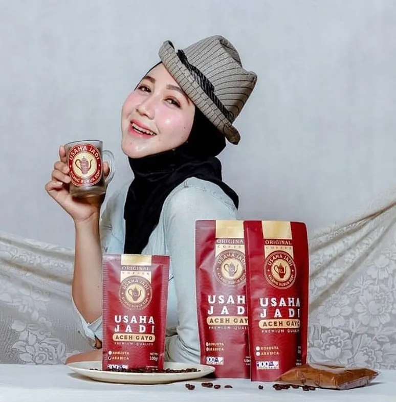 Perempuan berhijab dan bertopi memegang cangkir kaca berlogo Usaha Jadi, di depan tiga pouch kopi merah Aceh Gayo