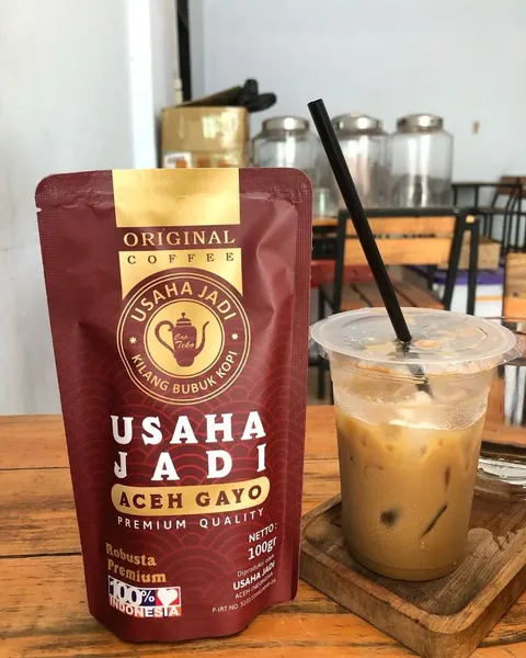Pouch merah marun Usaha Jadi Aceh Gayo Robusta Premium 100 g di samping es kopi susu bersedotan di meja kayu