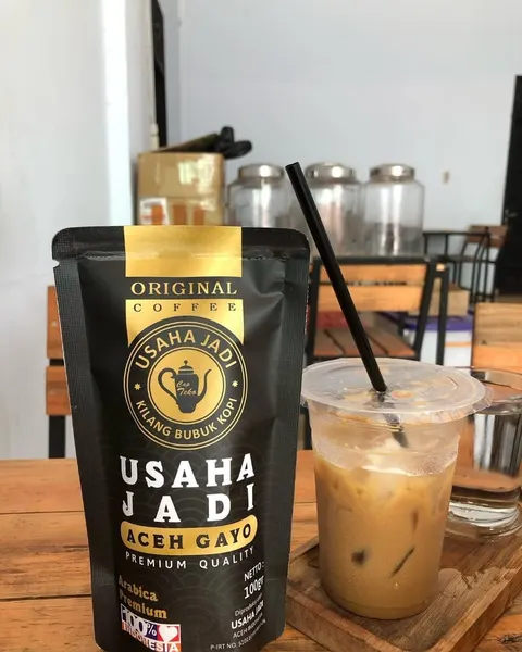 Pouch hitam Usaha Jadi Aceh Gayo Arabica Premium 100 g di samping es kopi susu bersedotan di meja kayu