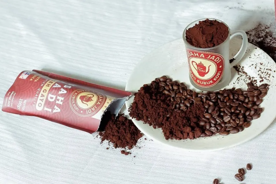 Pouch merah Usaha Jadi terbuka berisi bubuk kopi, cangkir penuh bubuk kopi, dan biji kopi di piring putih